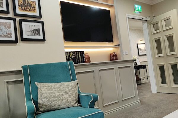 At JFW Cabinet Ltd, we take pride in delivering bespoke furniture solutions that enhance the functionality and aesthetics of any space. Our recent project for Kingsley Healthcare's new care home, Sycamore Heights in Bedford, is a testament to our commitment to quality and craftsmanship. Reception Desk We crafted an elegantly curved reception desk with solid surface tops with fronts designed to match the corridor wall panelling. This desk not only serves as a welcoming focal point but also seamlessly integrates with the overall design of the entrance area. Wall Panelling This traditional shaker-style wall panelling was produced in sections in our workshop, allowing for fast and efficient installation on site. This panelling was incorporated into the entrance lobby and the Quiet Lounge, adding a touch of classic elegance to these spaces. Signing-in Desk Located in the entrance lobby, the signing-in desk was manufactured to match the wall panelling and fit perfectly into an alcove. This design ensures discreet access to the gas meter while maintaining a cohesive look. Quiet Lounge Kitchenette The Quiet Lounge features a beautiful traditional kitchenette with solid surface tops and shaker-style doors. This kitchenette seamlessly ties in with the room's wall panelling, creating a harmonious and inviting space. Quiet Lounge TV Cabinet Designed to complement the kitchenette, the TV cabinet features a top section that houses LED lighting and a TV. This unit blends seamlessly with the wall panelling, enhancing the room's functionality and style. Cafe Unit The stylish cafe unit includes a drinks cooler and coffee machine, along with ample storage. This unit is designed to provide both functionality and a modern aesthetic. Quiet Room Bookcase An angled open shelf bookcase fits perfectly into the eaves of the second-floor Quiet Lounge, providing practical storage and a charming feature. TV Fireplace Bookcase This multifunctional cabinet incorporates an electric fire, television, cupboard units, and open shelving, adding warmth and versatility to the space. Alcove Unit A lovely feature in the Quiet Lounge, the alcove unit allows more light into the room, enhancing its ambiance. Staff Base Counters We installed four nurse base counters around the building to assist staff in providing the best possible care for the residents. We collaborated closely with HomeSmiths Ltd to produce furniture from their designs. Our team conducted site surveys to measure the space and then created working drawings for approval, and meticulously crafted each piece in our workshop before installation. Visit HomeSmiths Ltd homesmiths.co.uk to learn more about their designs. For more information about our bespoke furniture solutions, contact us at JFW Cabinet Ltd.