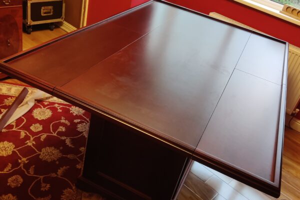 This bespoke gaming table was designed and crafted to deliver total functionality within a compact living space, without compromising on style. Positioned within a bay window, the brief was to create a table that could remain neatly tucked away for everyday use… Yet transform effortlessly into a full-sized gaming surface when required. The solution was a cleverly engineered drop-leaf design, allowing the table to sit compactly into the bay window area before expanding to a generous 6ft x 4ft playing surface. This provides ample space for board games, cards, and Warhammer type games, all while maintaining a clean and unobtrusive footprint when not in use. Mobility was a key consideration. The table is mounted on four heavy-duty locking castors, enabling it to be moved with ease and securely fixed in position once in place. This flexibility ensures the table can adapt to the room as needed, whether for entertaining or everyday living. Internally, the cabinet features bifold doors in front of adjustable shelves, offering practical storage for games and accessories while maintaining a refined exterior appearance. High-quality hardware, including folding table hinges and precision joinery, ensures smooth operation and long-term durability. Crafted using mahogany veneered MDF with solid mahogany mouldings, the table was finished with a stain and lacquer carefully selected to complement the client’s existing furniture. Traditional detailing, including decorative handles, ties the piece seamlessly into the room’s established aesthetic. The result is a highly versatile, beautifully crafted piece that balances practicality with timeless design a perfect example of bespoke furniture tailored to both space and lifestyle. If you’re considering a bespoke piece tailored to your space, explore more of our recent projects or get in touch to discuss your ideas. At JFW Cabinetmakers, we specialise in creating furniture that works beautifully for the way you live.